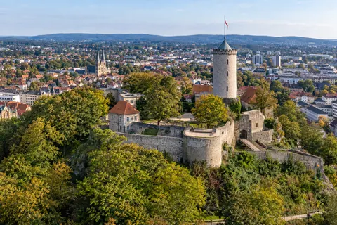Illustrative Stadtansicht von Bielefeld mit dem runden Turm der Sparrenburg auf einem bewaldeten Hügel.